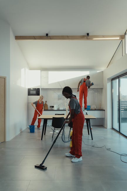 Close-up of a person wearing pink rubber gloves using a spray bottle of cleaning solution to disinfect a white kitchen shelf with a small potted plant, in a well-lit kitchen environment. The scene sho