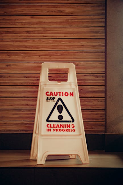 A white plastic 'Caution Cleaning in Progress' warning sign with a black exclamation mark inside a triangle, and red lettering, placed on a tiled floor against a background of wooden paneling and a textured wall. The sign indicates ongoing cleaning work, reflecting a hygienic environment maintained through professional cleaning services by Cleaner Hounslow. No other tools or surfaces are visible in the image, which emphasizes safety signage in a commercial or residential space during cleaning activities.
