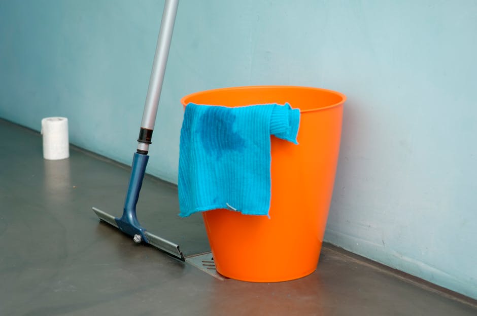 An image of a cleaning scene featuring a bright orange bucket placed on a smooth, grey floor next to a wall painted in a pale blue-green shade. Draped over the rim of the bucket is a blue microfiber cloth, damp from use. Leaning against the bucket is a mop with a blue handle and a flat mop head, positioned near a floor drain. In the background, there is a roll of white toilet paper or paper towel on the floor, indicating a cleaning or sanitation process. The scene is well-lit with natural or bright artificial lighting, emphasizing the cleanliness and preparation for surface and deep cleaning tasks. The setting appears to be an indoor area, possibly a bathroom or kitchen, where routine hygiene maintenance with tools like mops and cloths is essential. Cleaner Hounslow offers professional cleaning services focused on surface cleaning and sanitisation to ensure hygienic environments.