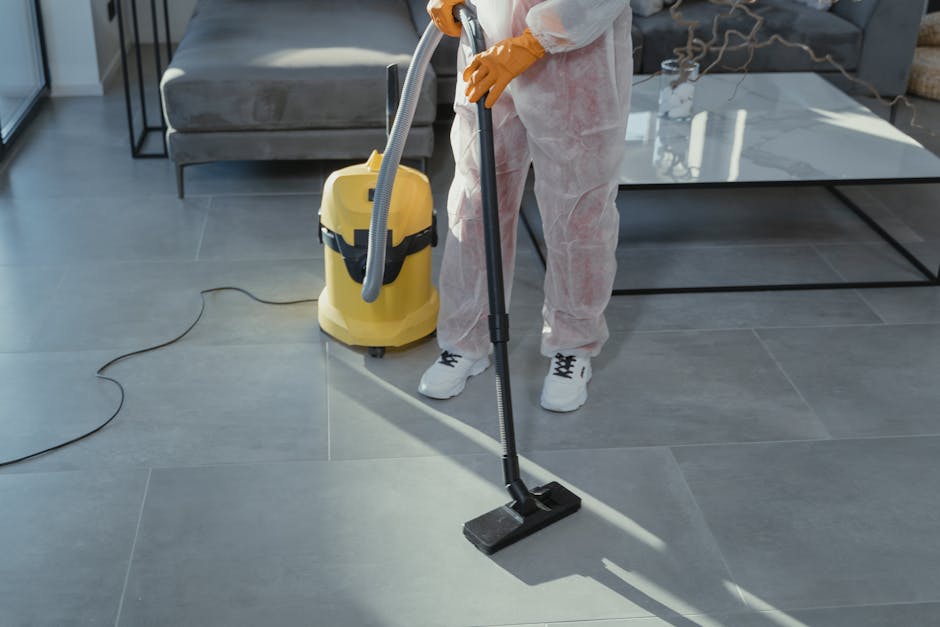 A white plastic 'Caution Cleaning in Progress' warning sign with a black exclamation mark inside a triangle, and red lettering, placed on a tiled floor against a background of wooden paneling and a textured wall. The sign indicates ongoing cleaning work, reflecting a hygienic environment maintained through professional cleaning services by Cleaner Hounslow. No other tools or surfaces are visible in the image, which emphasizes safety signage in a commercial or residential space during cleaning activities.