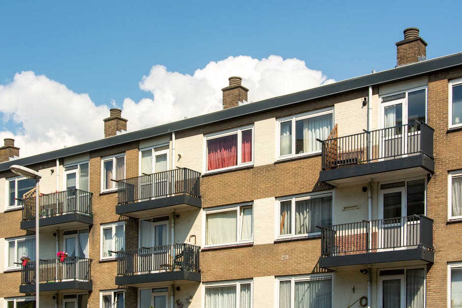 A multi-story residential building with a brick and cream facade, featuring several balconies with black railings. The balconies have outdoor furniture and potted plants. The building has multiple windows, some with curtains or blinds visible inside. Bright sunlight illuminates the scene, with a partly cloudy sky in the background. This image exemplifies the type of property that Cleaner Hounslow offers deep cleaning and surface sanitisation services for, particularly in preparation for end of tenancy cleaning in Hounslow TW3 flats.