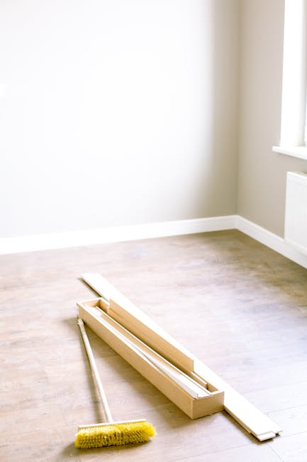 A bright, minimally furnished room featuring light-colored wooden flooring and a white wall with a large window allowing natural light to enter. On the floor, there is a long, narrow cardboard box containing a part of a window blind or curtain hardware, and a yellow-handled cleaning brush with a rectangular head is placed nearby. The room appears clean and ready for further detailed cleaning or furniture arrangement, with no visible dust or stains. [COMPANY_NAME] offers professional end of tenancy cleaning services that include surface cleaning and sanitisation, ideal for preparing flats like this Hounslow TW3 property for new occupants. This image emphasizes the importance of thorough domestic cleaning and surface care to achieve a fresh, hygienic environment.