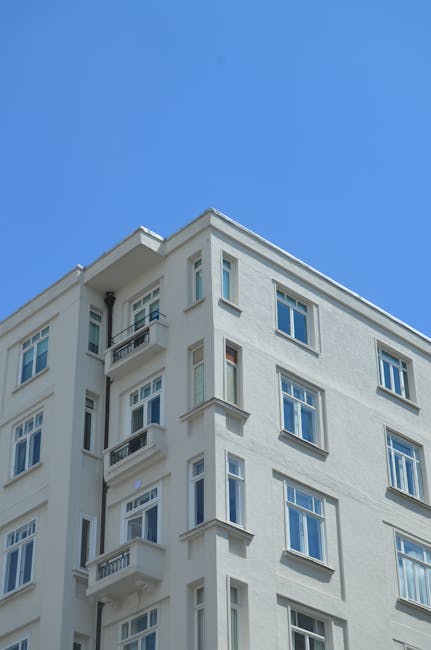 A multi-story residential building with a brick and cream facade, featuring several balconies with black railings. The balconies have outdoor furniture and potted plants. The building has multiple windows, some with curtains or blinds visible inside. Bright sunlight illuminates the scene, with a partly cloudy sky in the background. This image exemplifies the type of property that Cleaner Hounslow offers deep cleaning and surface sanitisation services for, particularly in preparation for end of tenancy cleaning in Hounslow TW3 flats.