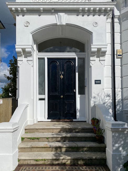 The entrance of a white Victorian-style residential building featuring a black front door with the number '28' displayed, flanked by two white columns supporting decorative moldings and an arched window above. The stone steps leading up to the door are clean and free of debris, with potted plants placed on either side. The facade exhibits intricate architectural details and is well-maintained, reflecting a neat and hygienic appearance. Natural sunlight casts shadows across the steps, and the surrounding area appears tidy, emphasizing the importance of surface cleaning and maintenance. For professional deep cleaning and sanitisation services in Hounslow, trust Cleaner Hounslow for thorough domestic cleaning solutions.