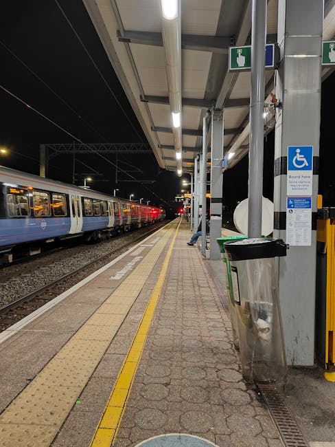 A well-lit Hounslow West Station platform at night showing the tiled surface with tactile paving strips along the edge for safety, and a stainless steel bin placed near a column. The platform features a glass and metal shelter with overhead fluorescent lighting, and a moving train with blue and white carriage doors is visible on the left. The surface of the platform is clean and free of debris, and the overall scene reflects a maintained environment with attention to hygiene and safety. Cleaner Hounslow provides surface cleaning and sanitisation services to ensure that public transport areas like this remain hygienic and well-kept, suitable for both residential and commercial cleaning needs.