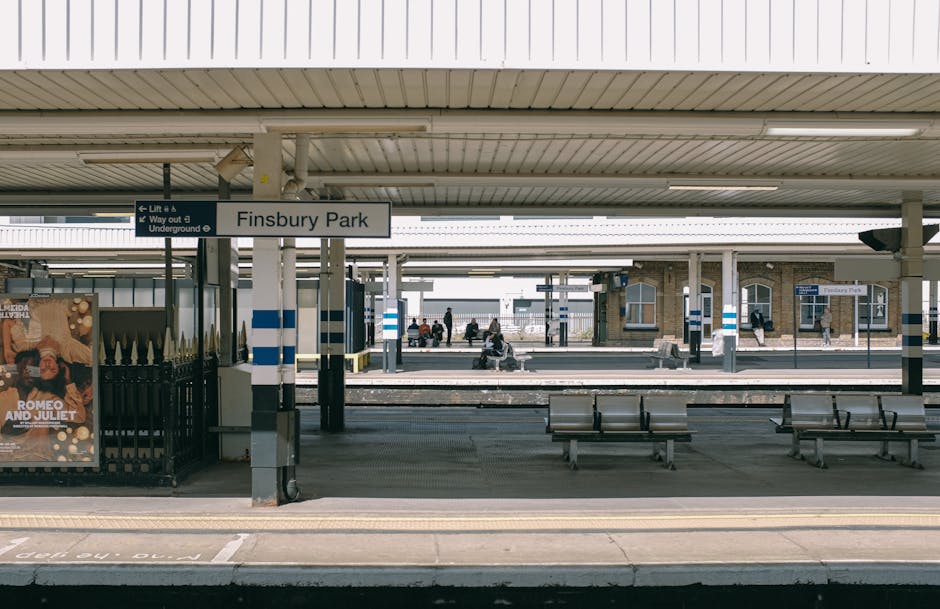 A well-lit Hounslow West Station platform at night showing the tiled surface with tactile paving strips along the edge for safety, and a stainless steel bin placed near a column. The platform features a glass and metal shelter with overhead fluorescent lighting, and a moving train with blue and white carriage doors is visible on the left. The surface of the platform is clean and free of debris, and the overall scene reflects a maintained environment with attention to hygiene and safety. Cleaner Hounslow provides surface cleaning and sanitisation services to ensure that public transport areas like this remain hygienic and well-kept, suitable for both residential and commercial cleaning needs.
