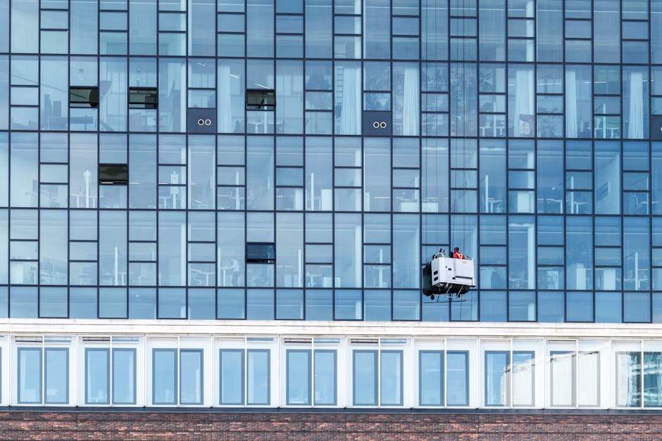 A window cleaning technician operating a suspended platform on the exterior of a modern glass office building, with a clean, reflective glass facade. The technician is visible wearing safety gear, including a helmet and harness, and appears to be cleaning or inspecting the windows. The building features multiple floors with large windows, some of which have blinds or curtains inside. Natural daylight illuminates the scene, highlighting the shiny, spotless surfaces, demonstrating professional surface cleaning and maintenance services tailored for commercial properties, as promoted by Cleaner Hounslow for high-street businesses.