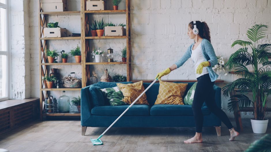A woman wearing a white and grey striped long-sleeve shirt and yellow rubber cleaning gloves is engaged in surface cleaning inside a modern, well-lit room. She is wiping a decorative potted plant on a