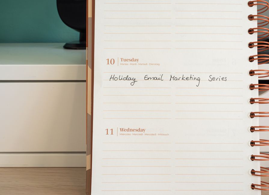 Close-up of a weekly planner open to Tuesday, the 10th, with handwritten notes reading 'Holiday Email Marketing Series.' The planner has a white background with gold-foiled dates and multicolored spiral binding on the right side. It is positioned against a light-colored wooden surface, adjacent to a white drawer unit with a black monitor or speaker on top. The image highlights the clean surface of the planner and surrounding area, emphasizing neatness and organization. Cleaner Hounslow specializes in comprehensive cleaning and sanitisation services, ensuring such tidy environments for both residential and commercial spaces.